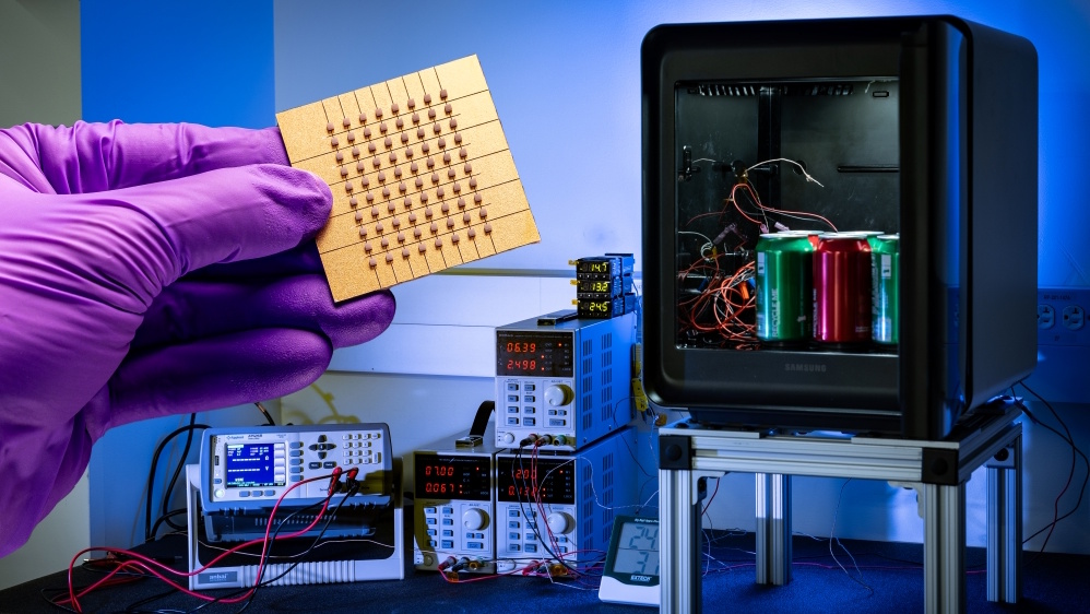 A Samsung and Johns Hopkins APL Peltier effect cooling device being held up by a hand with a lab glove, with various electronic gubbins going on in the background